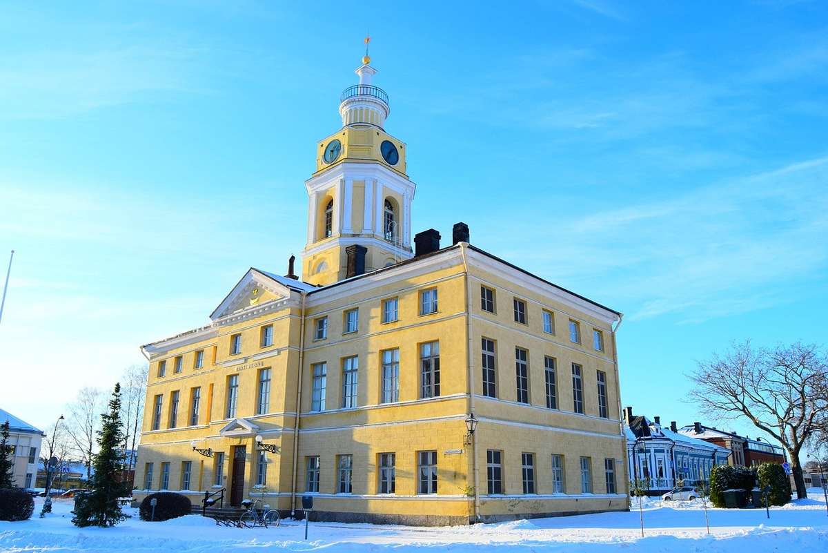 Bâtiment jaune à toits pentus, flèche centrale avec horloge, façade blanche, colonnes, devant un ciel bleu Bâtiment jaune à toits pentus, flèche centrale avec horloge, façade blanche, colonnes, devant un ciel bleu